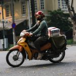 Entregador usando capacete vermelho pilota uma motocicleta por uma rua urbana enquanto transporta um pacote.