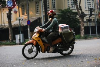 Entregador usando capacete vermelho pilota uma motocicleta por uma rua urbana enquanto transporta um pacote.
