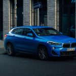 SUV BMW azul estacionado em uma rua urbana molhada, cercado por edifícios modernos.
