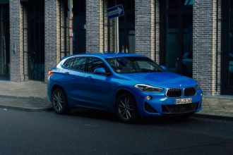 SUV BMW azul estacionado em uma rua urbana molhada, cercado por edifícios modernos.