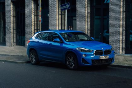 SUV BMW azul estacionado em uma rua urbana molhada, cercado por edifícios modernos.