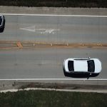 Vista aérea de dois carros brancos trafegando por uma rodovia com marcações direcionais.