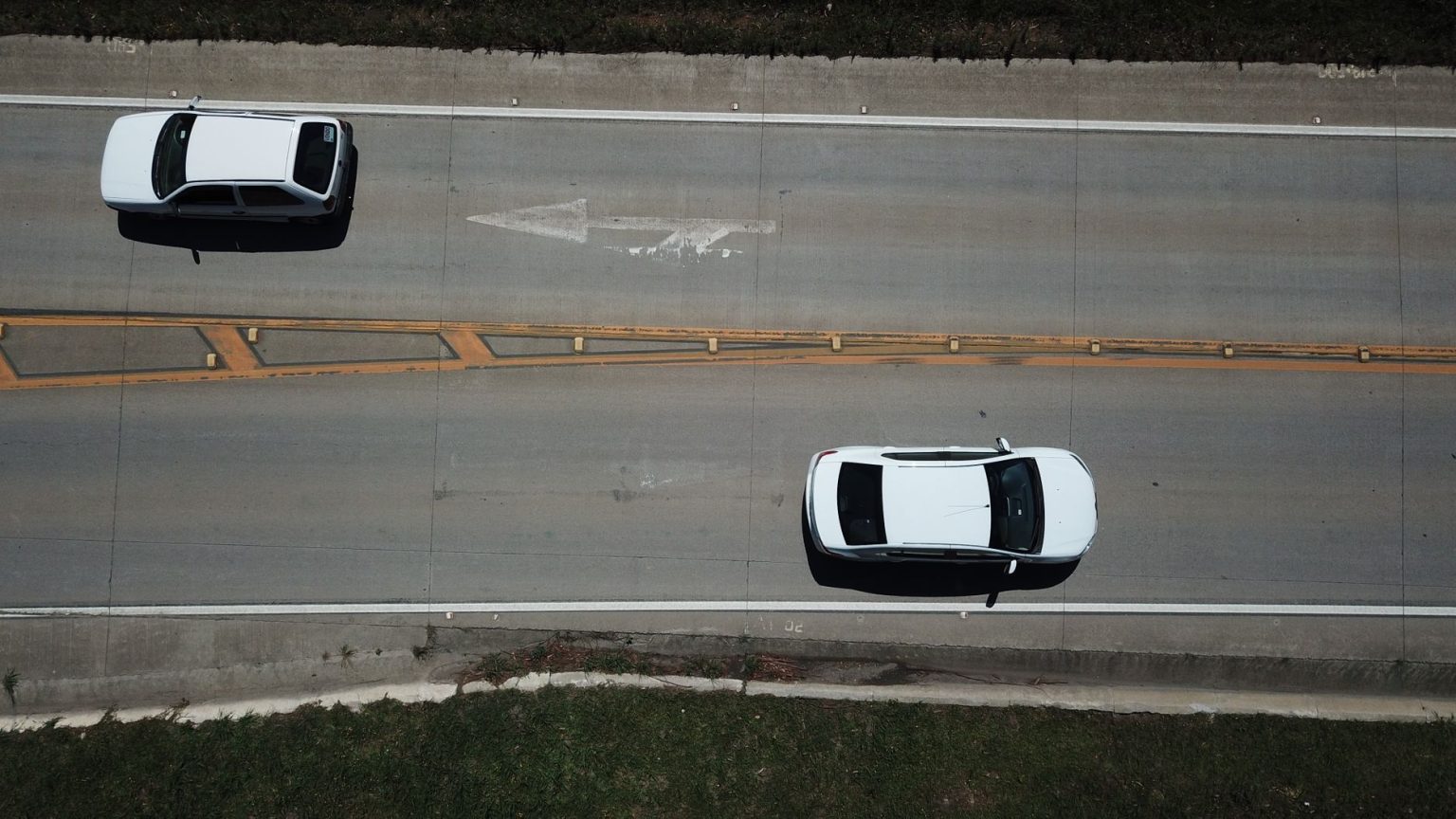 Vista aérea de dois carros brancos trafegando por uma rodovia com marcações direcionais.