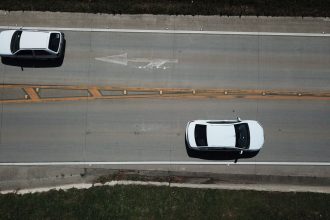 Vista aérea de dois carros brancos trafegando por uma rodovia com marcações direcionais.