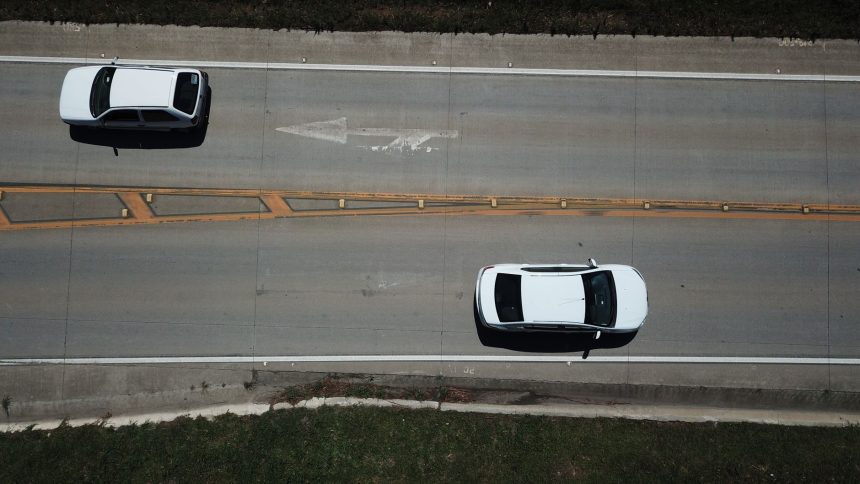 Vista aérea de dois carros brancos trafegando por uma rodovia com marcações direcionais.